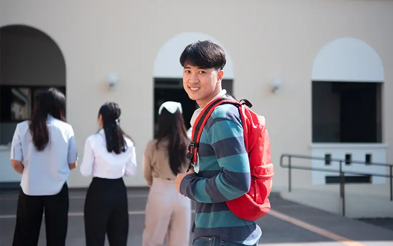 Một sinh viên đại học đang mỉm cười với chiếc ba lô màu đỏ đang đứng trên khuôn viên trường, tự tin nhìn lại trong khi những sinh viên khác đang ở phía sau.