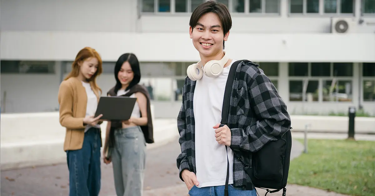 Một nam sinh viên vui vẻ đứng trên khuôn viên trường đại học với tai nghe đeo quanh cổ và ba lô, trong khi hai nữ sinh viên đang học trên máy tính xách tay ở phía sau