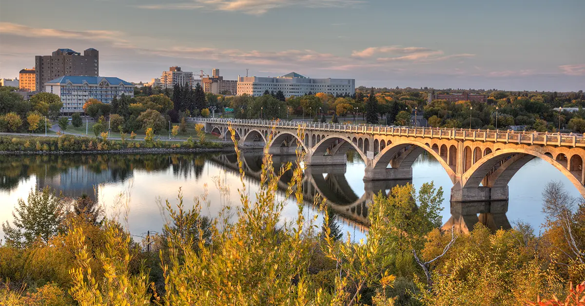 Hình ảnh ngang của đường chân trời Saskatoon, Saskatchewan lúc chạng vạng. Hình ảnh được chụp vào đầu mùa thu với tán lá nhiều màu hiện rõ ở phía trước. Khách sạn, chung cư và bệnh viện hiện rõ ở phía xa của Sông Nam Saskatchewan. Cầu Broadway có thể được nhìn thấy ở phía bên phải của hình ảnh cùng với hình ảnh phản chiếu gần như hoàn hảo trên mặt nước.